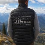Man standing with his back to the camera on a mountain overlook, wearing a black quilted vest with an alpine scene and German text on the back.