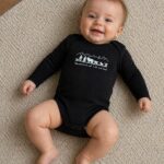 Smiling baby lying on a beige carpet, wearing a black onesie with a white graphic of mountains and animals.