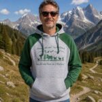 Man wearing a gray and green hoodie and sunglasses stands on a mountain trail with snow-capped peaks in the background, smiling at the camera.