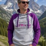 Man in sunglasses stands outdoors in a mountain landscape, wearing a gray hoodie with purple sleeves and front pocket pockets.