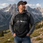 Man stands with hands in pockets on a rocky mountain trail, wearing a black and gray hooded sweatshirt and a cap, with snow-capped peaks in the background.