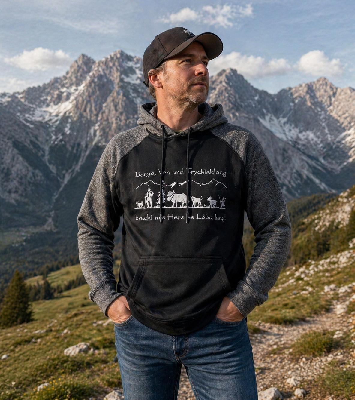 WhatsApp Image 2026-04-30 at 1352386 | fil - magic Stickerei Berther Man stands with hands in pockets on a rocky mountain trail, wearing a black and gray hooded sweatshirt and a cap, with snow-capped peaks in the background.