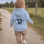 Back view of a toddler walking along a gravel path, wearing a light blue hoodie with a circular cow-themed design on the back.