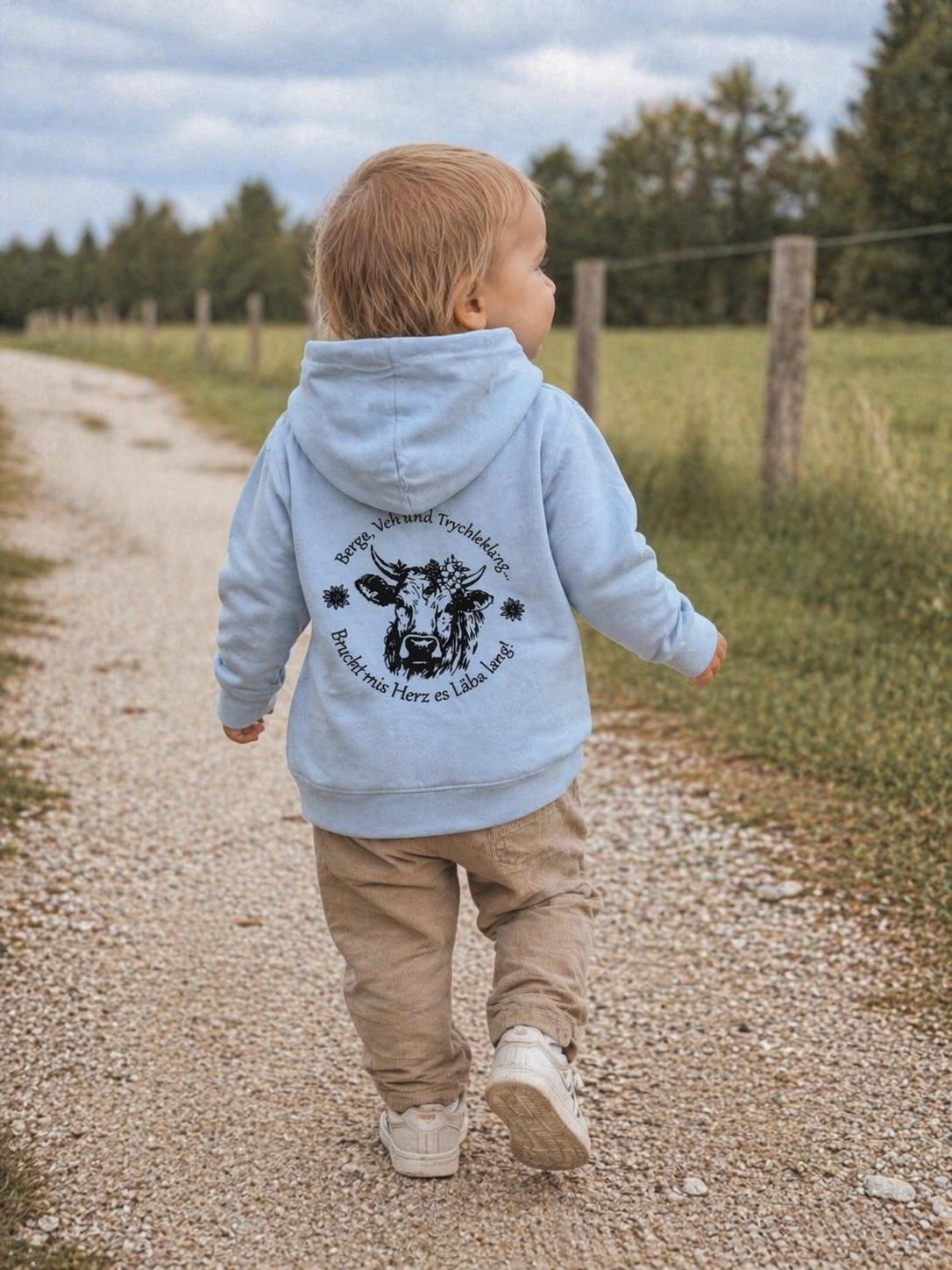 WhatsApp Image 2026-04-30 at 135301 | fil - magic Stickerei Berther Back view of a toddler walking along a gravel path, wearing a light blue hoodie with a circular cow-themed design on the back.