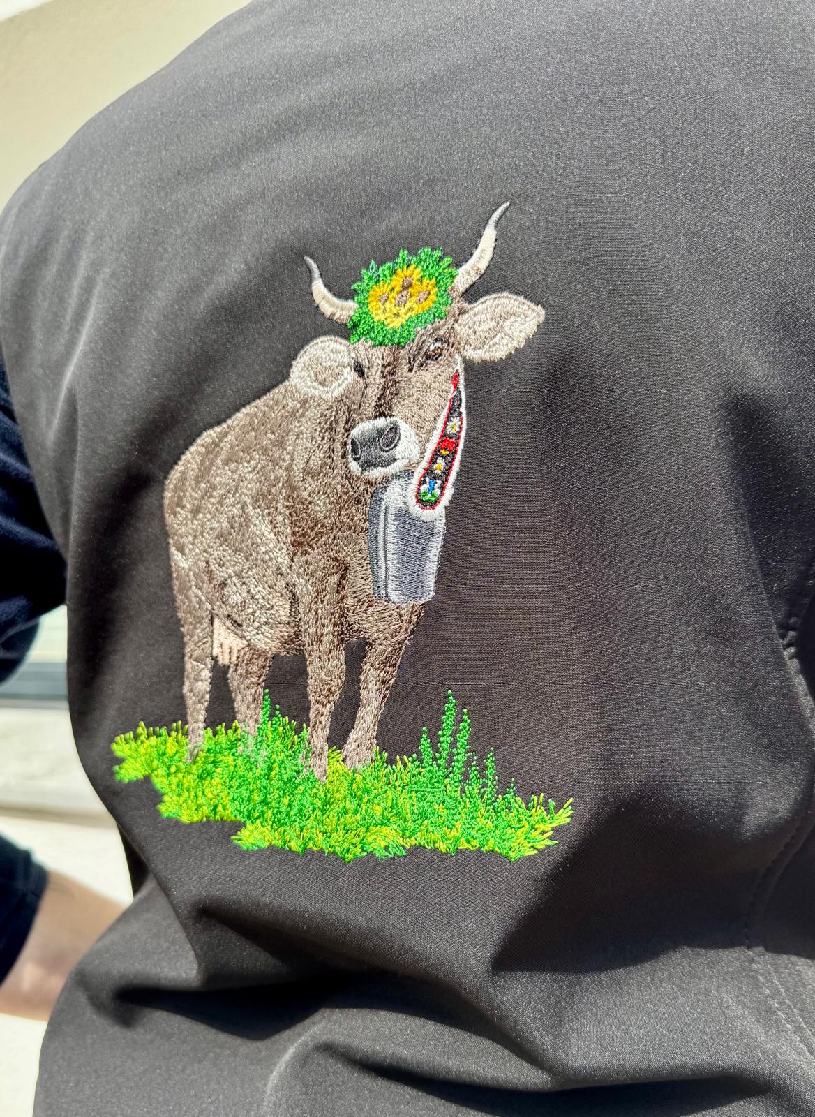WhatsApp Image 2026-04-30 at 1353531 | fil - magic Stickerei Berther Embroidered cow with a green floral crown on its head, standing on grass, on black fabric.