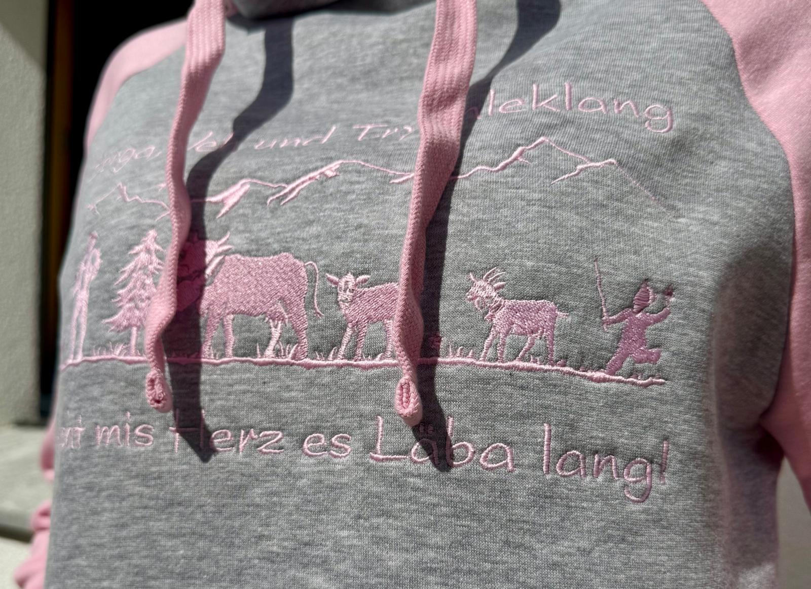 WhatsApp Image 2026-04-30 at 1353534 | fil - magic Stickerei Berther Gray hoodie with pink embroidery showing a mountain landscape, trees, a line of animals, and a small child raising a stick, plus cursive text along the bottom in pink thread.