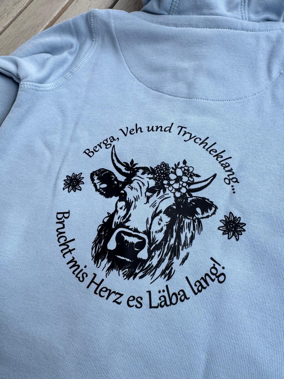 WhatsApp Image 2026-04-30 at 1353541 | fil - magic Stickerei Berther Light blue hoodie with a circular black illustration of a cow adorned with flowers, surrounded by German text.