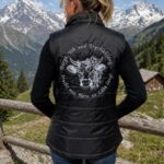 Woman standing outdoors with back to camera, wearing a black padded vest with a white bull graphic and circular German text, in a mountainous landscape.