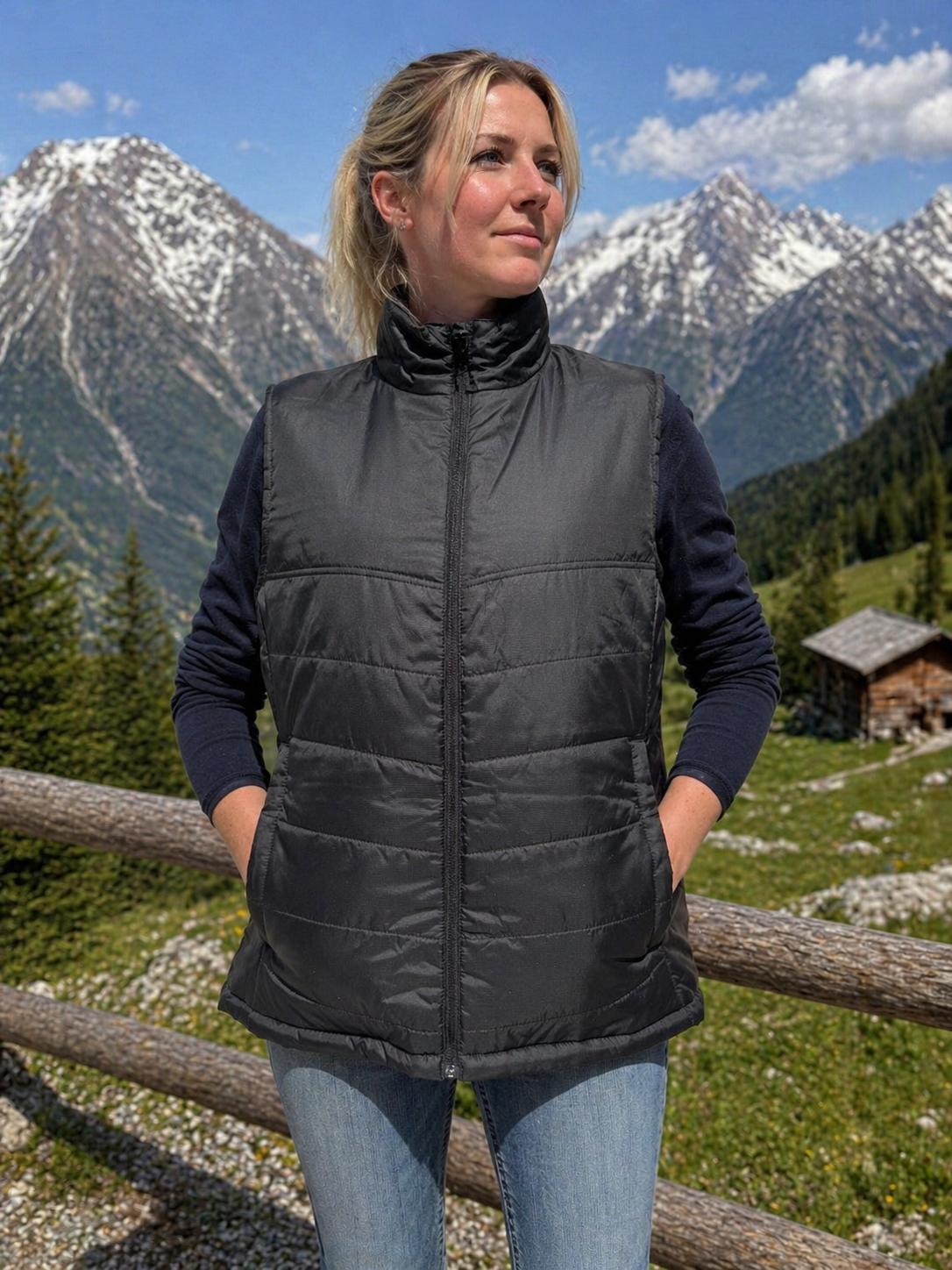 WhatsApp Image 2026-05-20 at 1114401 | fil - magic Stickerei Berther Woman standing outdoors in a black puffer vest over a dark long-sleeve shirt, hands in pockets, against snow-capped mountains and a wooden fence.