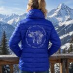Person seen from behind wearing a bright blue puffer jacket with a circular white graphic on the back, standing at a wooden railing with snow-covered mountains in the background.