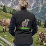 Back view of a person in a black vest with an embroidered cow design, standing in a green alpine meadow with mountains in the background