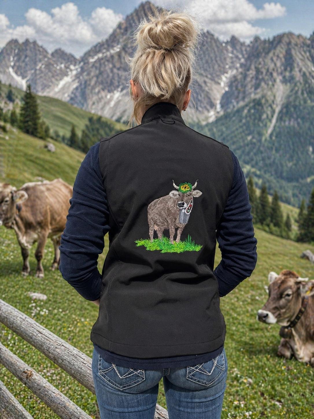 WhatsApp Image 2026-05-20 at 1114404 | fil - magic Stickerei Berther Back view of a person in a black vest with an embroidered cow design, standing in a green alpine meadow with mountains in the background