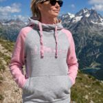 Woman in a gray and pink hoodie standing on a rocky mountain trail, wearing black sunglasses with snow-capped peaks in the background.