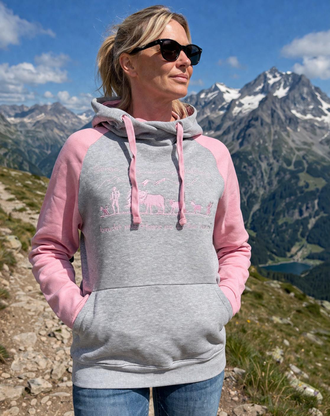 WhatsApp Image 2026-05-20 at 1114406 | fil - magic Stickerei Berther Woman in a gray and pink hoodie standing on a rocky mountain trail, wearing black sunglasses with snow-capped peaks in the background.