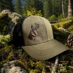 Olive green trucker hat with an embroidered llama resting on mossy rocks in a pine forest with mountains in the background