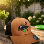 Brown trucker cap with black embroidered dog and green leaf on the front panel, outdoors on a wooden table in a sunlit garden shop