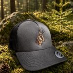 Black trucker cap with an embroidered deer head resting on moss in a sunlit forest.