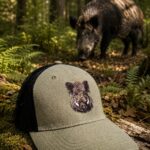 Olive-green trucker cap with a boar head embroidery on a mossy log in a forest, with a wild boar in the background.