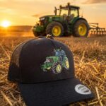 Black trucker cap with a green tractor embroidery in the foreground on a straw-strewn field at sunset; a tractor is visible in the distance.
