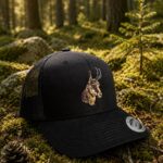 Black trucker cap with a tan embroidered antelope head sits on moss in a sunlit forest.