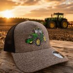 Grey mesh trucker hat with a green tractor embroidery, placed on a weathered wooden table at sunset, with a tractor in the field behind it.