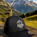 WhatsApp Image 2026-08-22 at 1728077 | fil - magic Stickerei Berther Black trucker cap with a Schwingen emblem rests on a weathered wooden surface, with snow-capped alpine mountains in the background.