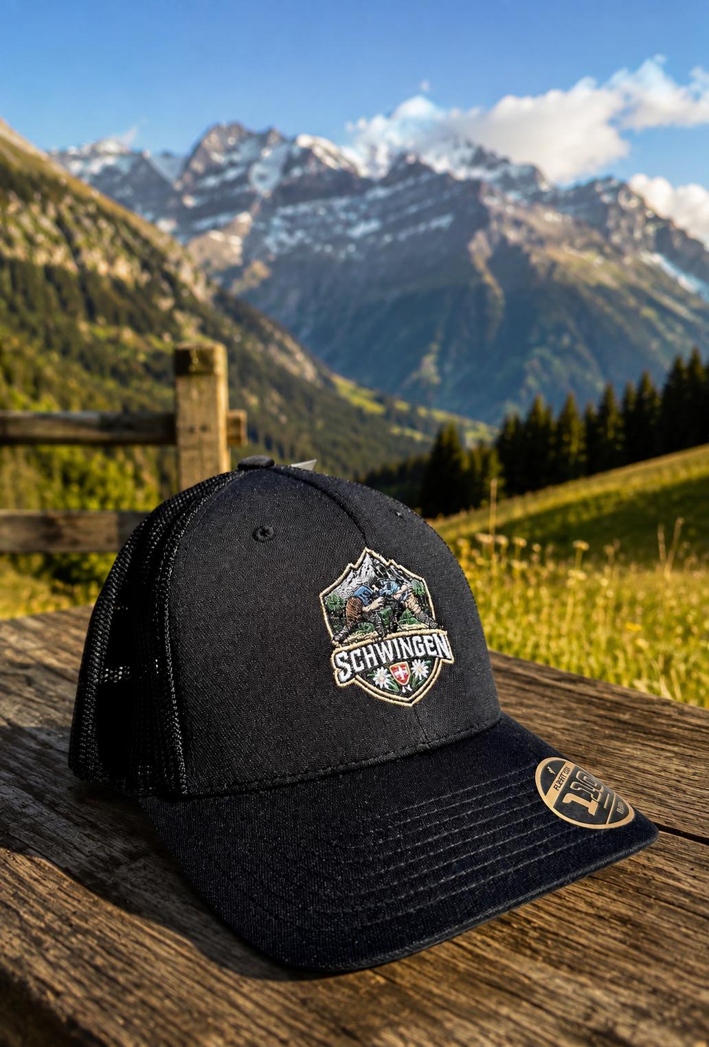WhatsApp Image 2026-08-22 at 1728077 | fil - magic Stickerei Berther Black trucker cap with a Schwingen emblem rests on a weathered wooden surface, with snow-capped alpine mountains in the background.