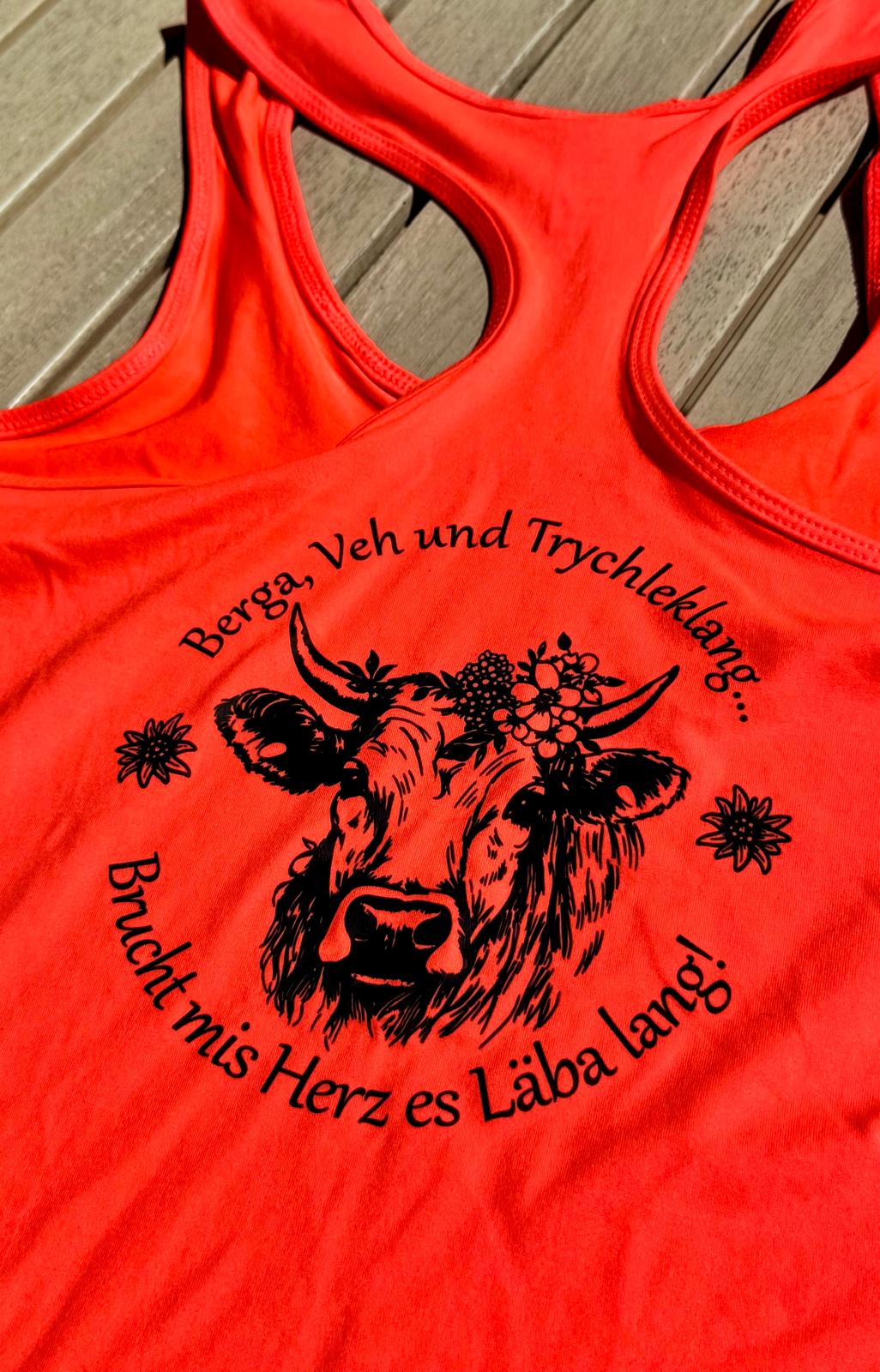 WhatsApp Image 2026-08-22 at 172909 | fil - magic Stickerei Berther Bright orange tank top laid on a wooden surface, featuring a circular black graphic of a cow's head with a flower crown and German slogan around it.